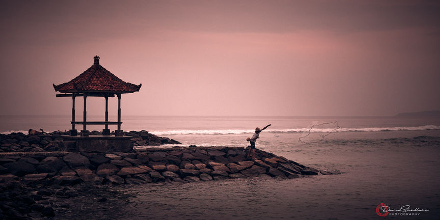 An epic image of a fisherman throwing his net off the edge of a rock jetty with a soft pink sky and an asian outbuilding.