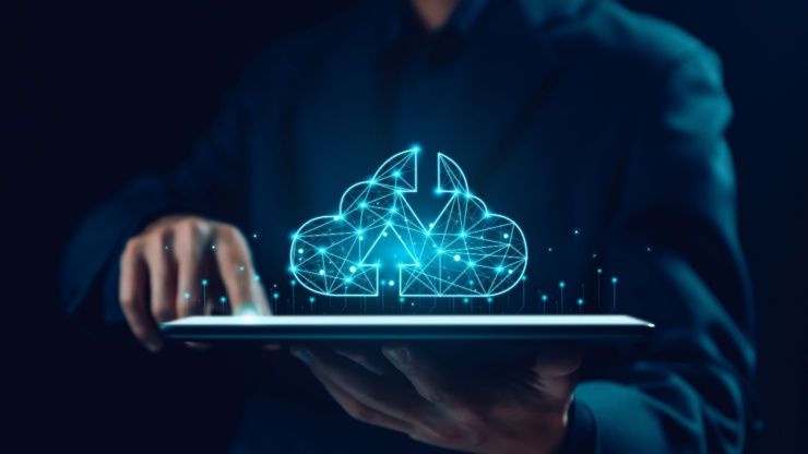 A person in a dark suit holds a tablet with a glowing blue cloud and circuit pattern above it, suggesting digital technology or data.