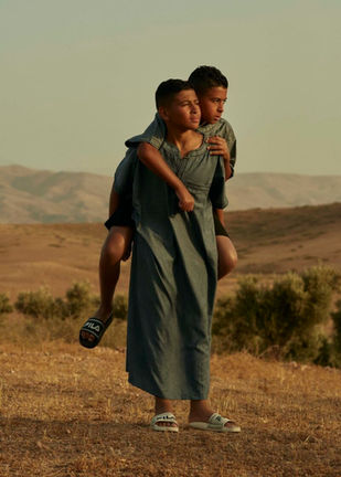 Een Marokaanse jongen draagt een andere jongen op zijn rug. Ze dragen allebei een Djellaba en lopen in een heuvelachtig droog landschap