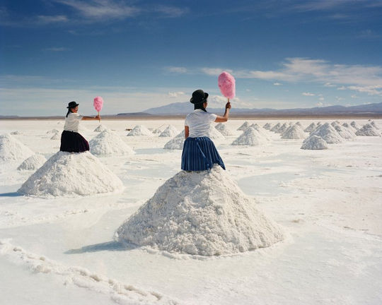 Zoutvlakte in Bolivia met heuveltjes zout. Op twee ervan zit een vrouw in Boliviaanse kledrdracht met een roze suikerspin in de hand.