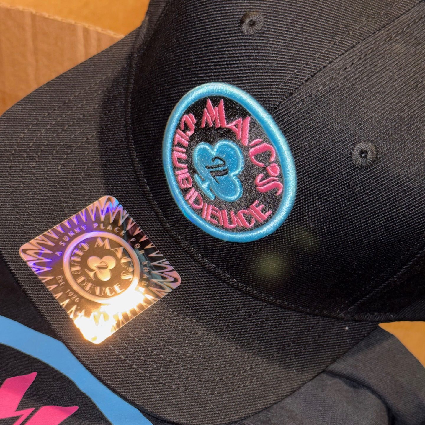 Close-up of the Mac's Club Deuce logo in Miami Vice pink and blue on a snapback baseball cap.
