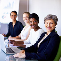 Four smiling businesswomen at a meeting with laptops and a whiteboard in background