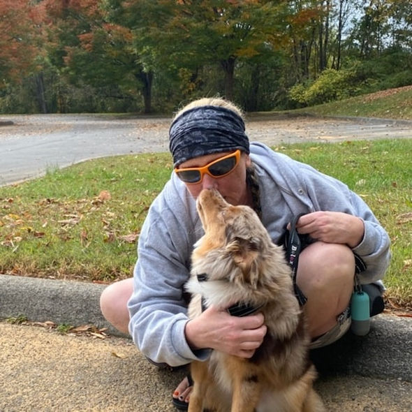 Crouching person wearing bandana lovingly nuzzles their friendly dog by the road.