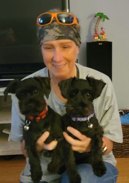 Person wearing hat and sunglasses holding two adorable black puppies.