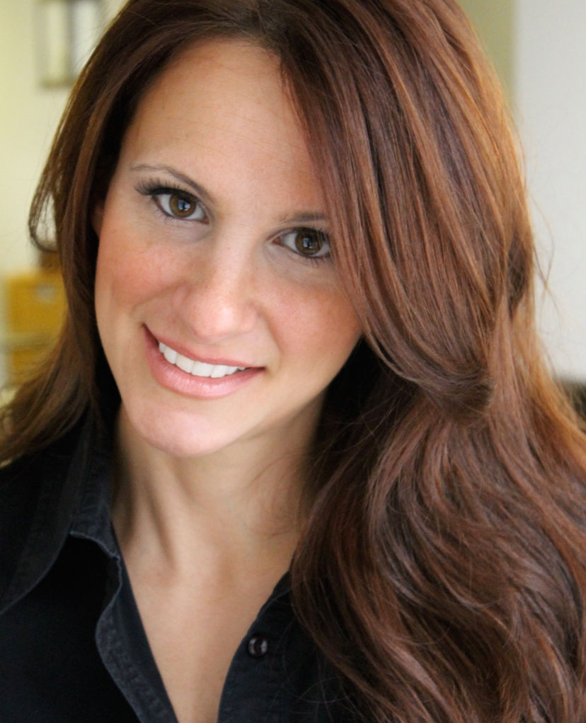 Smiling woman with long brown hair wearing a black shirt. Neutral indoor background. Bright and approachable expression.