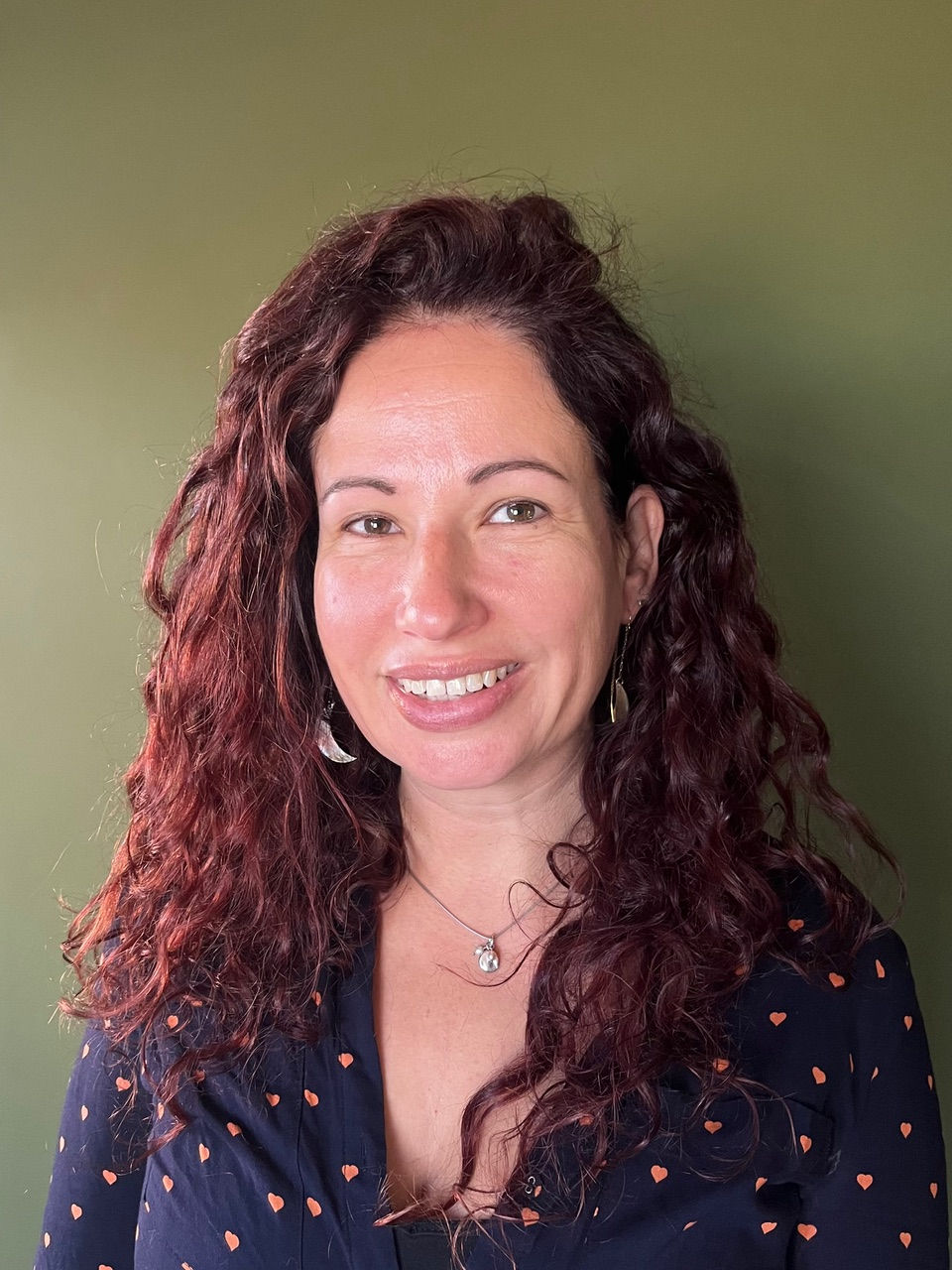 Smiling woman with wavy hair, wearing earrings and a heart-patterned blouse, stands against a plain olive-green background.