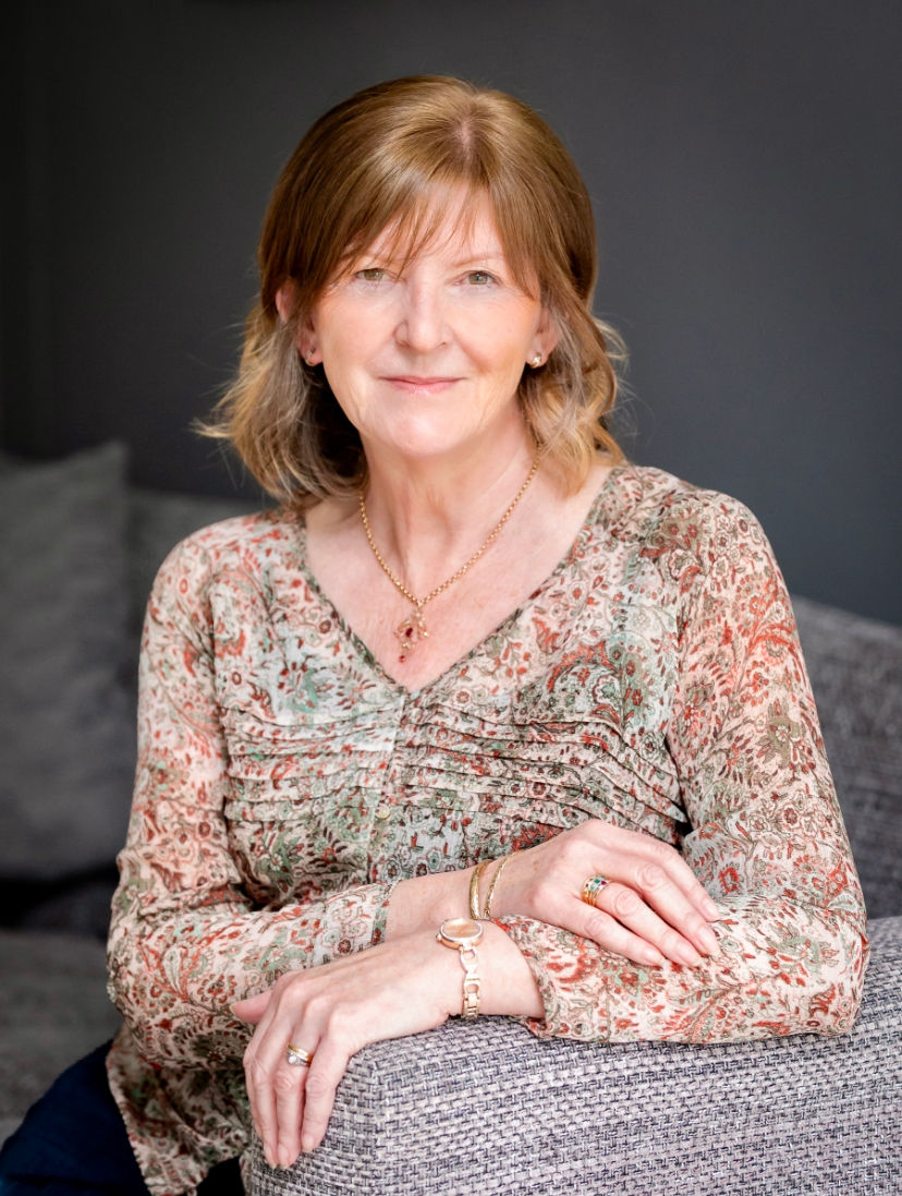 White woman with dark blonde hair seated with arms resting on gray upholstered arm chair