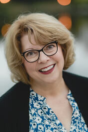 Smiling woman with glasses, wearing a blue floral top and black cardigan. Soft-focus lights in the blurred background create a warm mood.