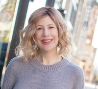 Smiling woman with wavy blonde hair in a gray sweater stands by a window with a blurred city background.