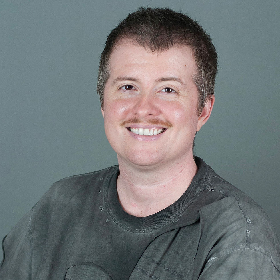 Smiling person with short hair and a mustache, wearing a gray shirt, against a plain gray background. Mood is cheerful.