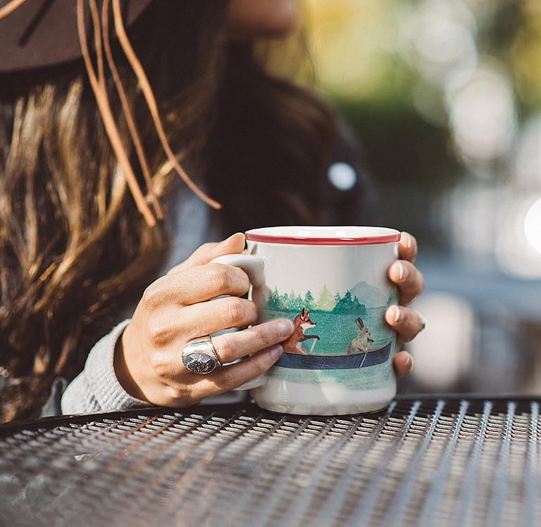 Hands holding white coffee mug with red stripe and painted image of bear paddling red canoe