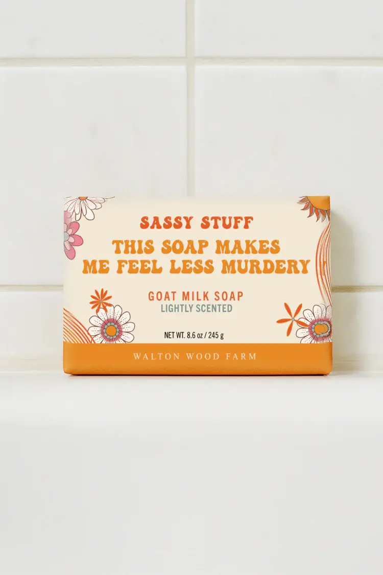 Orange & yellow paper wrapped bar soap labelled This Soap Makes Me Fell Less Murdery bar soap in white tiled bathroom