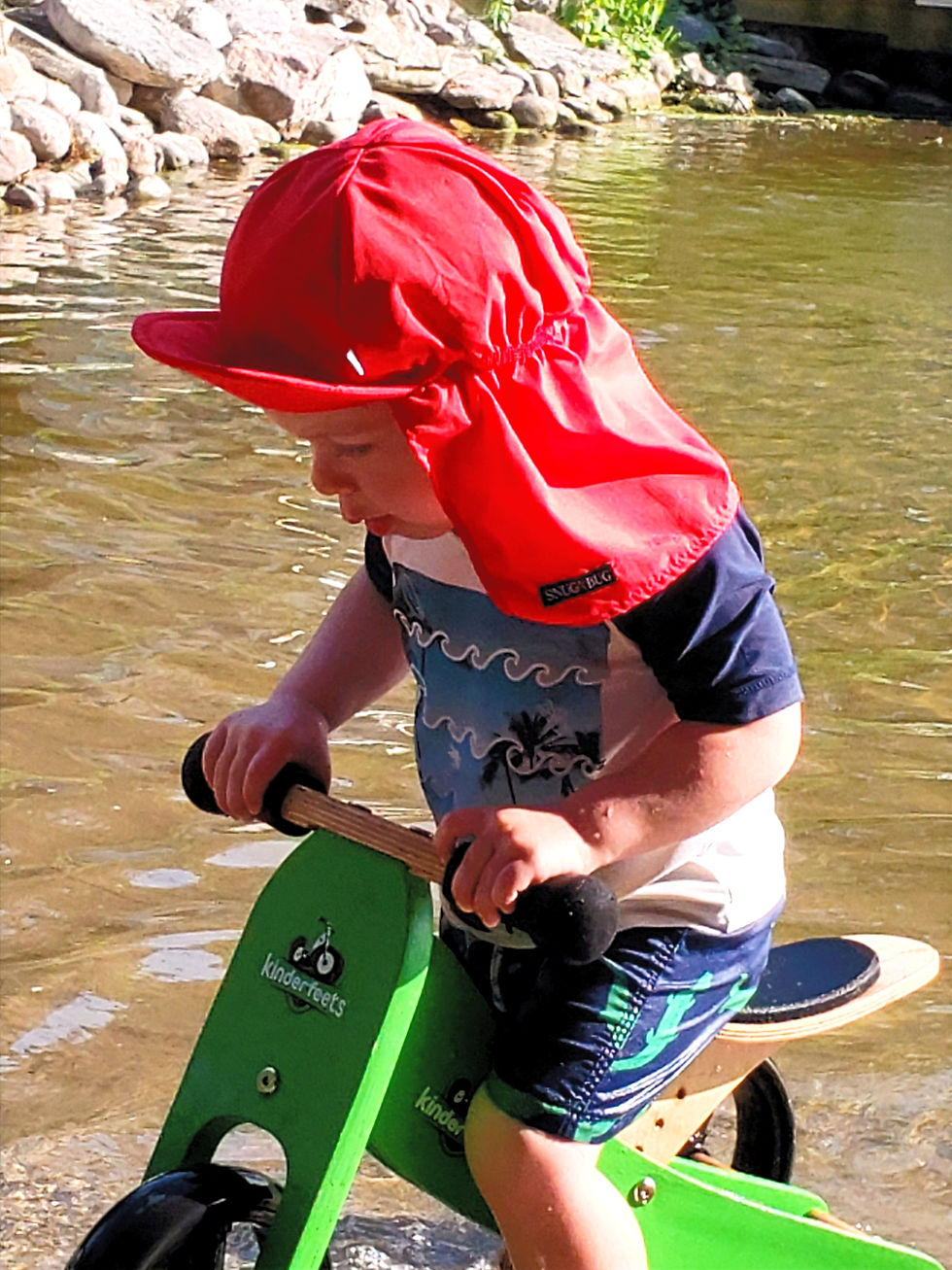 Baby boy on a trike in shallow water wearing a red cap style sun hat with peaked brim