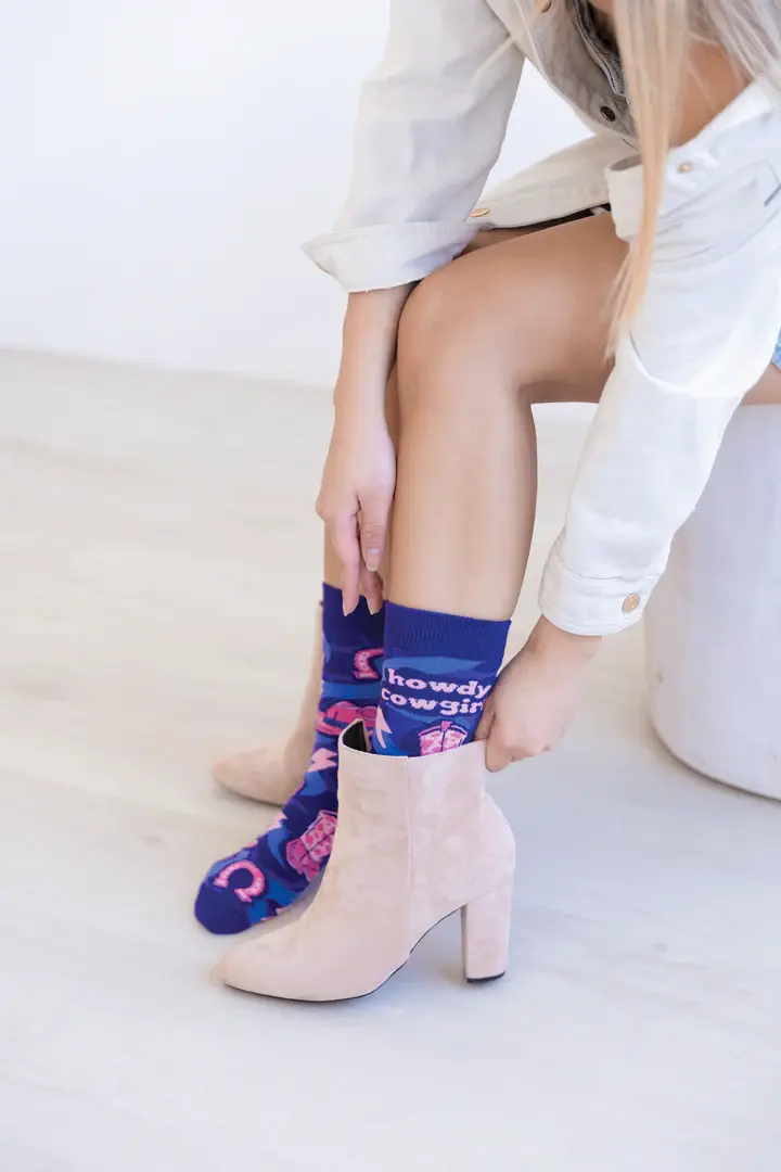 Seated model wearing navy socks with pink & blue print of cowgirl boots & cacti, text on side, 'Howdy Cowgirl'