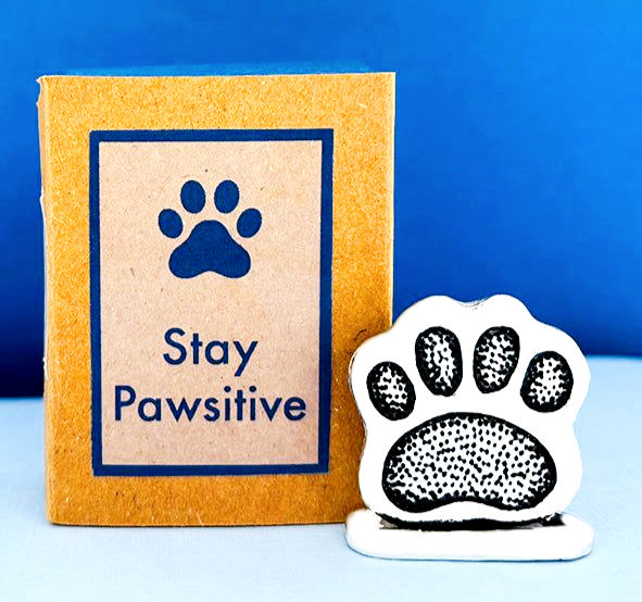 A Basic Spirit paper matchbook displaying "Stay Pawsitive" next to a textured pewter paw print miniature at Tickled Pink.