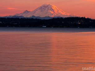 A cinematic view of Mount Rainier reflecting soft pink sunset light over the calm waters of the Pacific Northwest coastline.