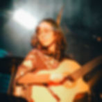 Mulher tocando violão em palco, com óculos e roupa estampada