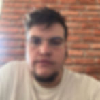 Homem de óculos usando camiseta branca frente a parede de tijolos