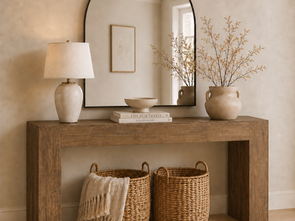 Warm minimalist entryway console table styled with an arched mirror, ceramic table lamp, stacked coffee table books, a catch-all bowl, textured vase with branches, and woven baskets underneath.
