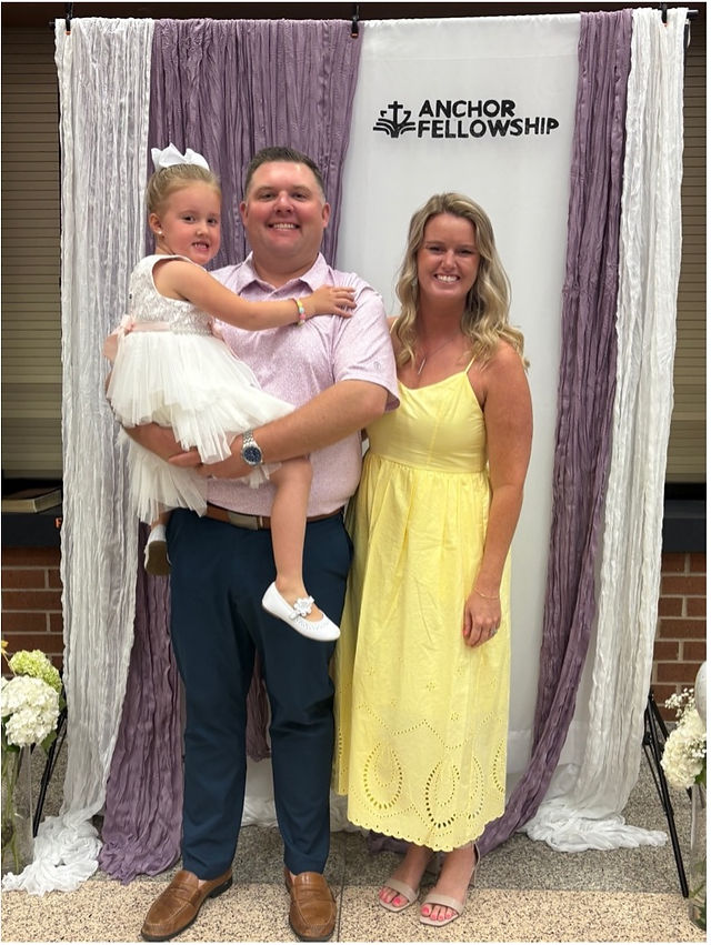 Family posing happily; man in pink shirt holding child in white dress; woman in yellow dress. "Anchor Fellowship" sign. Elegant backdrop.