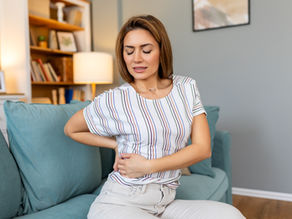 Person holding stomach in pain, wearing a white shirt with colorful stripes. Seated on a light blue sofa with a lamp in the background.