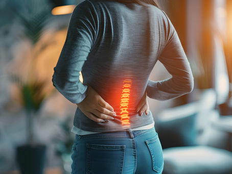 Person holding their lower back in discomfort, glowing spine overlay. Wearing gray shirt and jeans, indoor setting with soft lighting.
