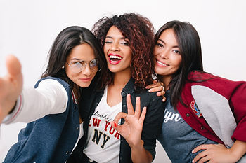 Three women pose cheerfully against a white background. One makes a 'peace' gesture. They're dressed casually; one wears aviator glasses.