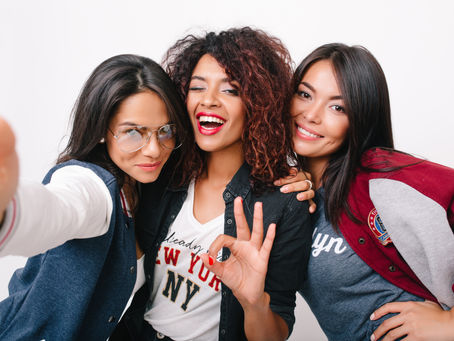 Three women pose cheerfully against a white background. One makes a 'peace' gesture. They're dressed casually; one wears aviator glasses.