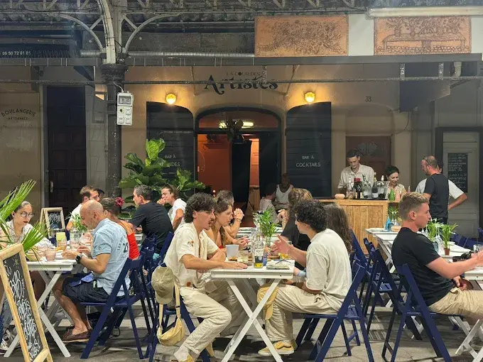 Busy outdoor terrace at La Cour des Artistes, Antibes at night. Guests seated at white folding tables with blue chairs under a covered arcade. Restaurant signage visible above the entrance. Street visible in the background.