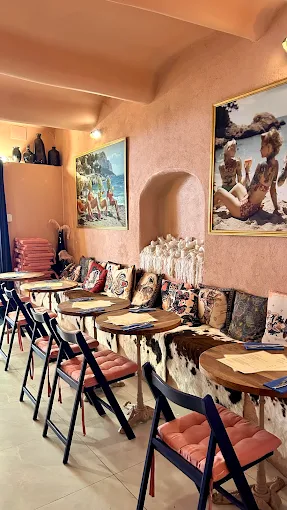 Dining area inside La Cour des Artistes with a long upholstered banquette seat in cowhide fabric, small round wooden tables, terracotta plaster walls, and large oil paintings. Warm, layered bohemian atmosphere.