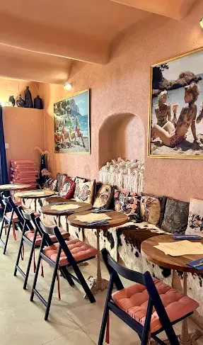 Dining area inside La Cour des Artistes with a long upholstered banquette seat in cowhide fabric, small round wooden tables, terracotta plaster walls, and large oil paintings. Warm, layered bohemian atmosphere.