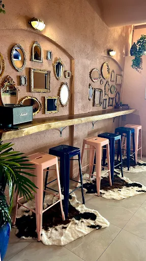 Interior detail of La Cour des Artistes showing a wall of mismatched vintage gold-framed mirrors above a live-edge wooden shelf. Pink and blue metal bar stools in the foreground, cowhide rug on the floor, tropical plant to the left.