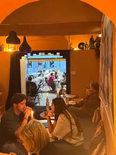 Intimate interior room at La Cour des Artistes lit by a single custom black ceramic pendant light by Naaya Studio. Low ceiling, warm candlelight, guests seated at close tables. View through an arched doorway to the outdoor terrace beyond.
