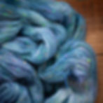 A pile of delicate haze yarn on a rustic wooden background. The yarn is a blend of soft blues and purples with bright specks of green and pink throughout the yarn.