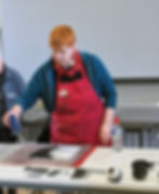 A woman in a red apron and blue sweater brushes felt on a table as other felting projects sit on the table nearby. She appears to be teaching a group felting techniques.