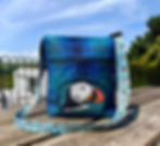 A dark blue, teal and dark green handbag outdoors ona wooden picnic table with a puffin head embroidered on the outside