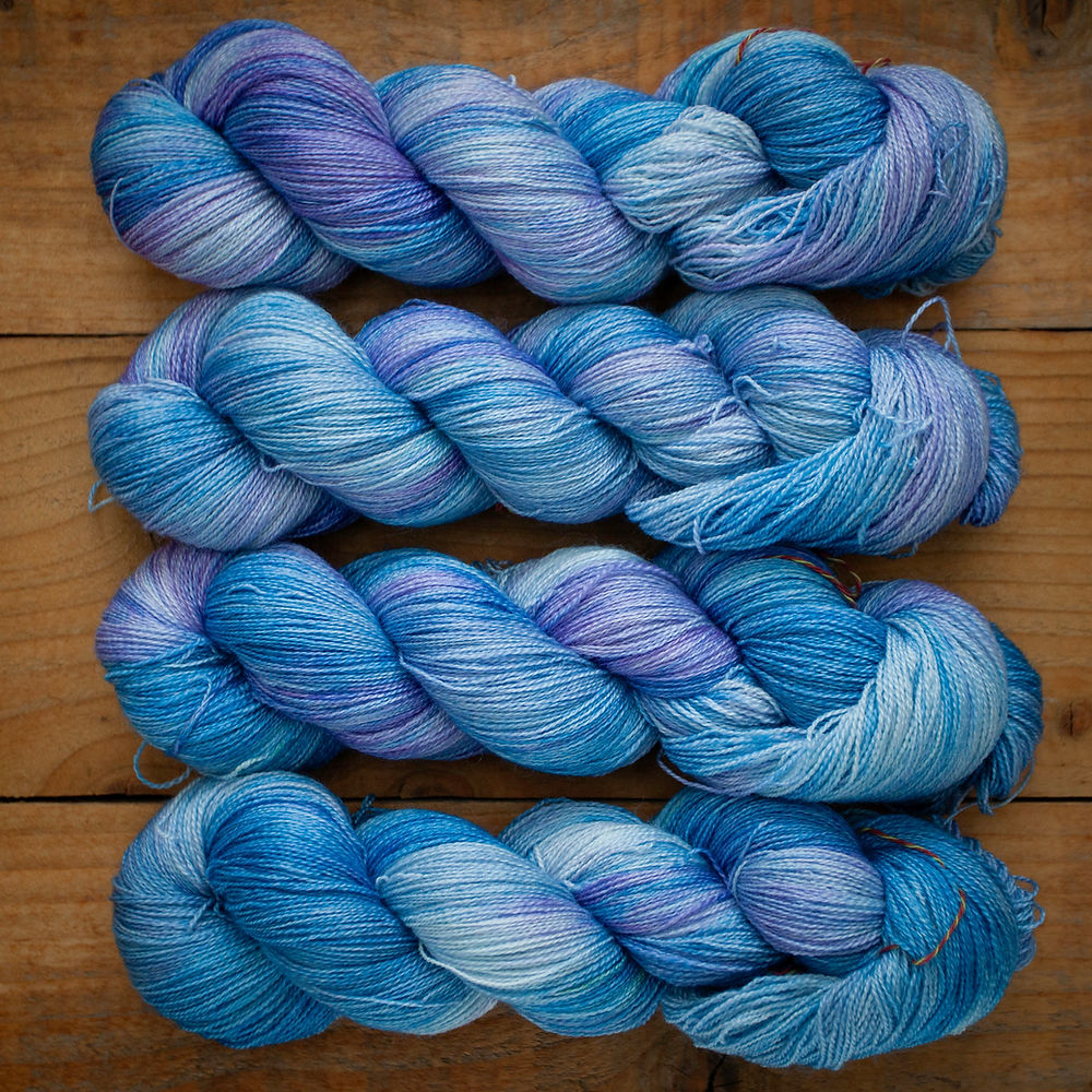 Four skeins of yarn on a rustic wooden background. The yarn is a blend of teals, light blues, and light purple.