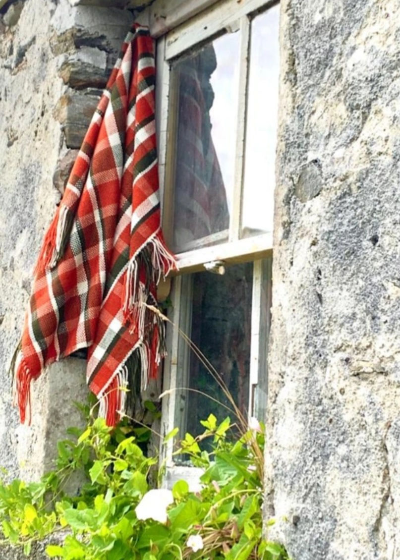 A woven checkered blanket in reds, whites and greys hangs from the window of a stone cottage with some green foliage in the foreground