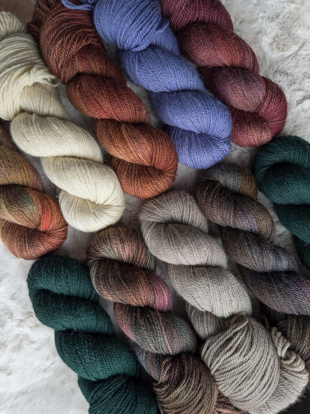 Ten skeins of yarn in various colours arranged neatly at an angle on a soft cream-coloured blanket, the yarns vary in colour from blues, reds, cream, greys and blacks and more