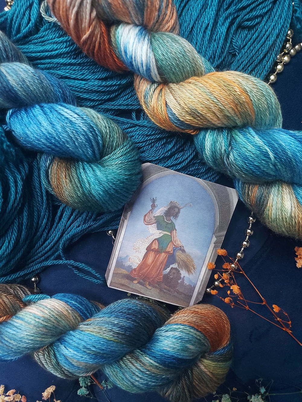 Three skeins of yarn in blues, browns creams and tans on a blue background surrounding a photo, taken from a 17thc image of a Black woman representing the continent of Africa she is wearing the same coloured garments and holding sheaf of wheat. This colourway is called Maternal Rainforest by Republic of Yarnia.