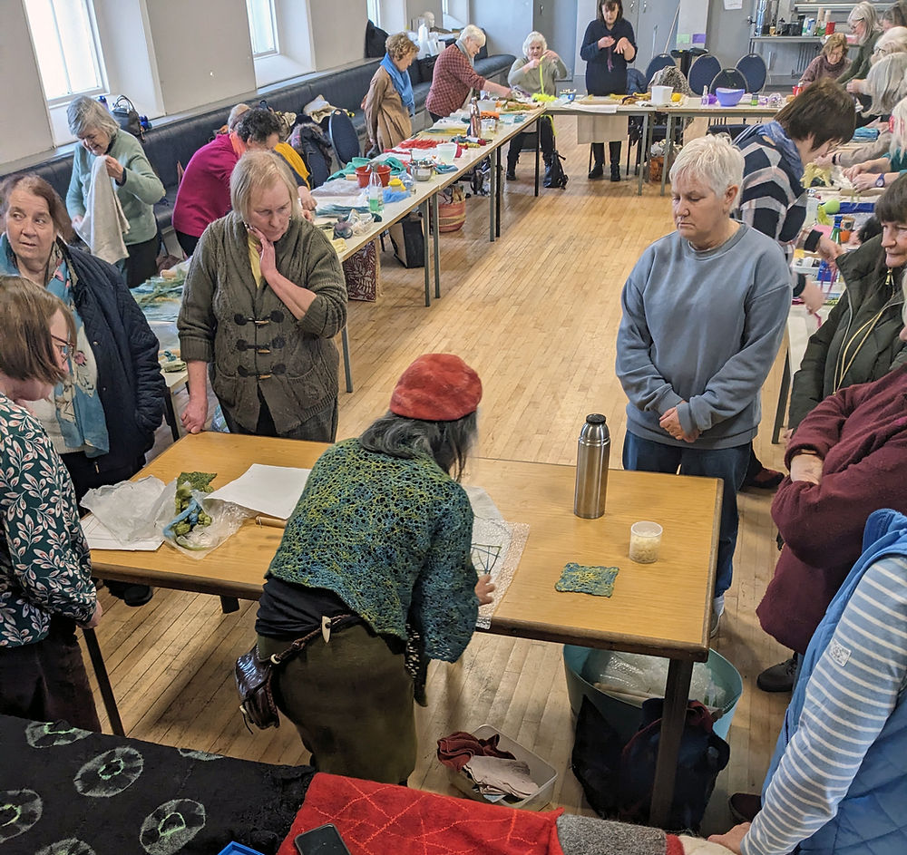 A group of about 20 women stand near tables in a room as some gather around a women in a green sweater leans over a table, working on a felting pattern.