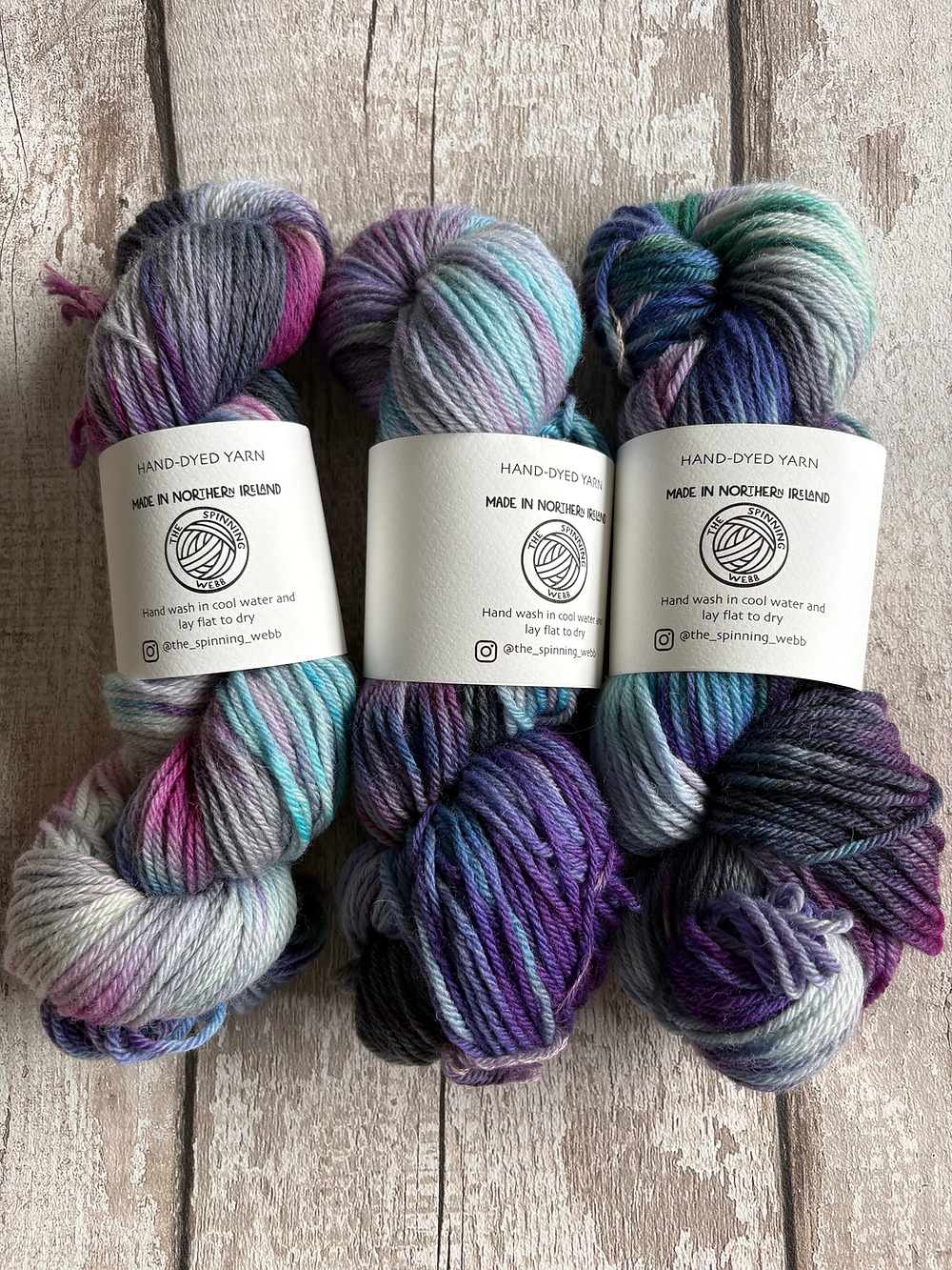 Three skeins of yarn in mixed blues, purples, greys and fushia with labels wrapped around them on a rustic, weathered wooden background.