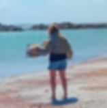 A woman stands on a windswept sandy beach in front of the water with rocky outcroppings in the distance. She has her back to us and is wrapped in a grey and light yellow striped blanket under a sunny sky