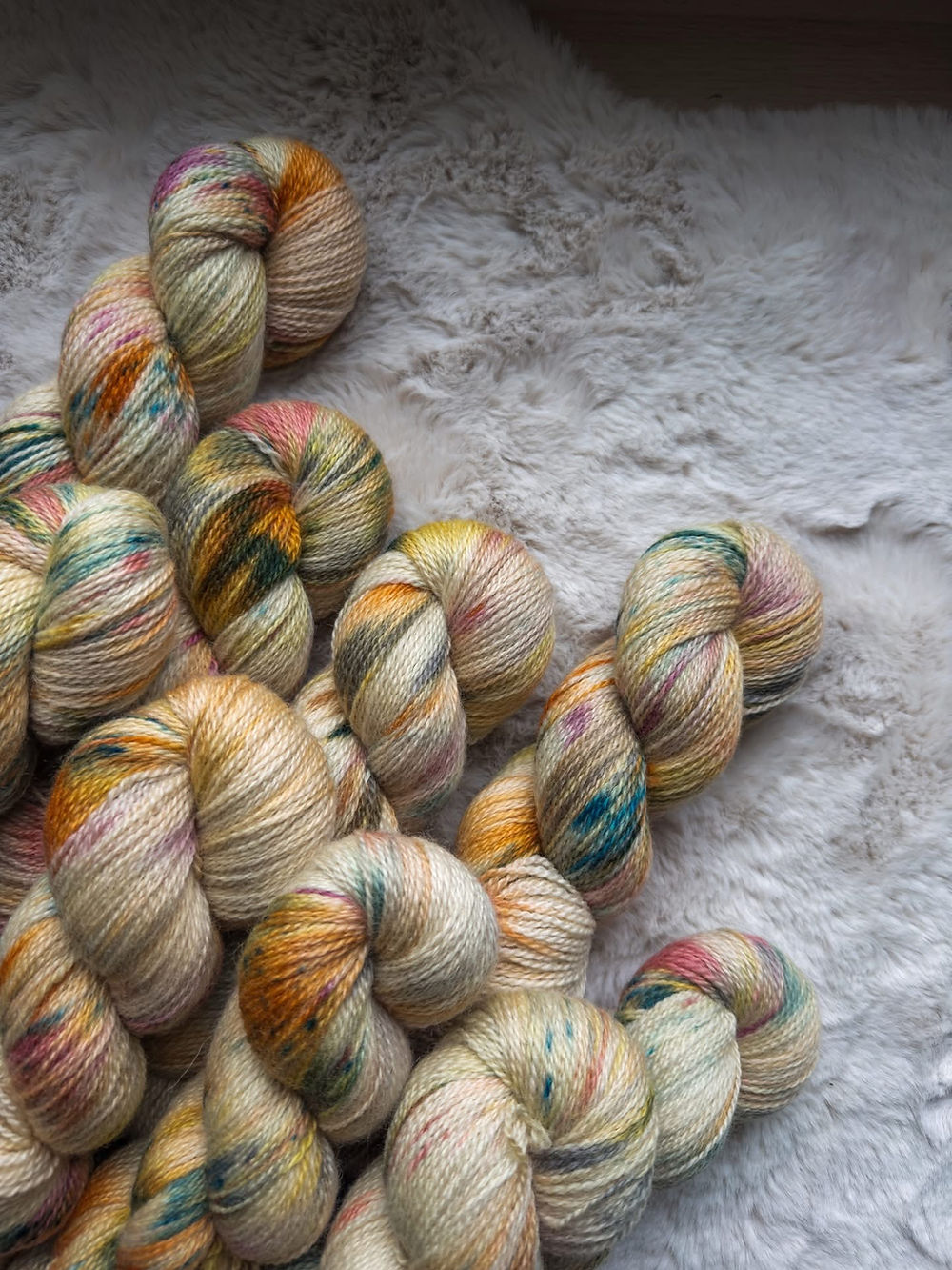 Eight skeins of yarn, photographed at an angle on a soft cream-coloured blanket. The yarn is a light tan with accents of orange, green and yellow.