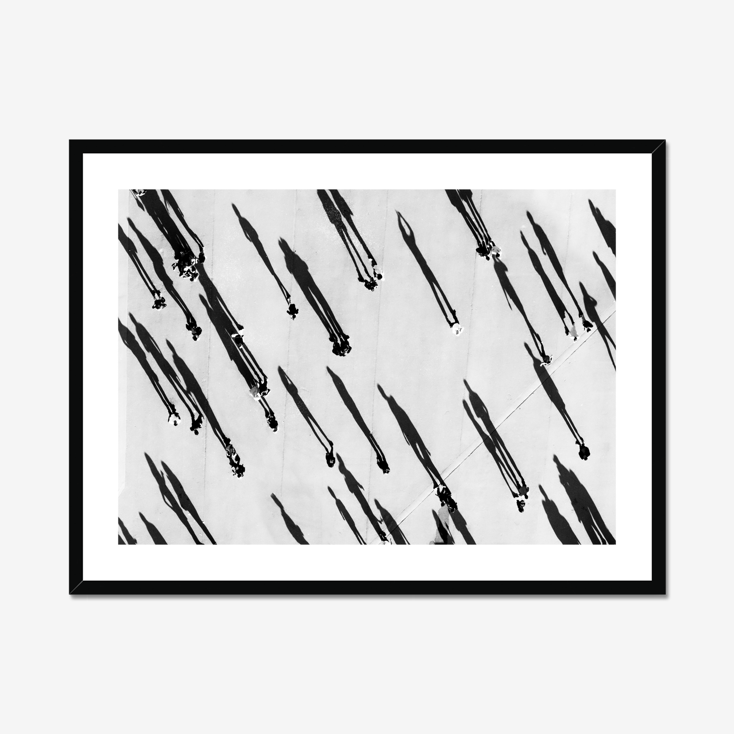 High contrast, fine art black and white photo of people from above, casting long diagonal shadows. Black frame with white mat