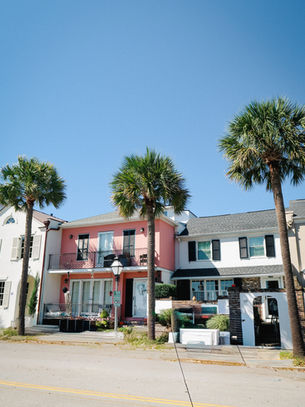 Colorful houses with balconies, flanked by palm trees under a clear blue sky. Pink and white facades create a vibrant, sunny street scene.