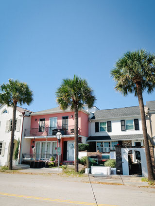 Colorful houses with balconies, flanked by palm trees under a clear blue sky. Pink and white facades create a vibrant, sunny street scene.