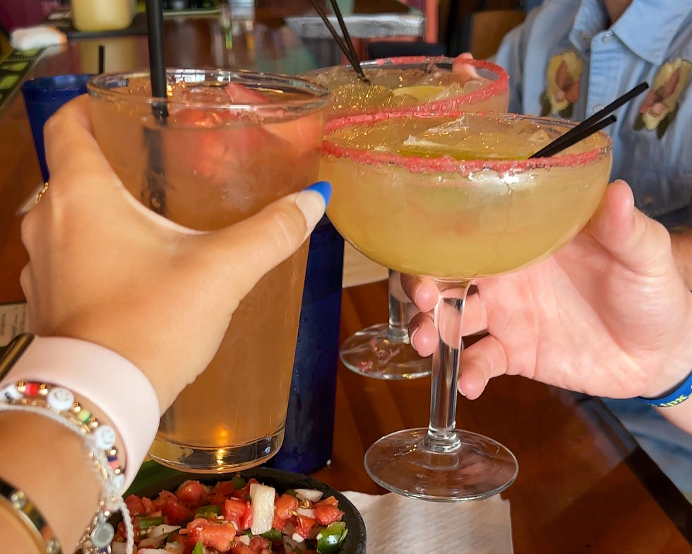 Hands clinking drinks, including margaritas with red sugar rims, over a table in a lively setting. Background features a blue shirt and food.