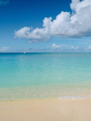 Calm beach with turquoise water and clear blue sky, white clouds in the background. Sandy shore in the foreground, creating a serene scene.