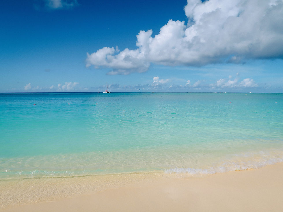 Calm beach with turquoise water and clear blue sky, white clouds in the background. Sandy shore in the foreground, creating a serene scene.